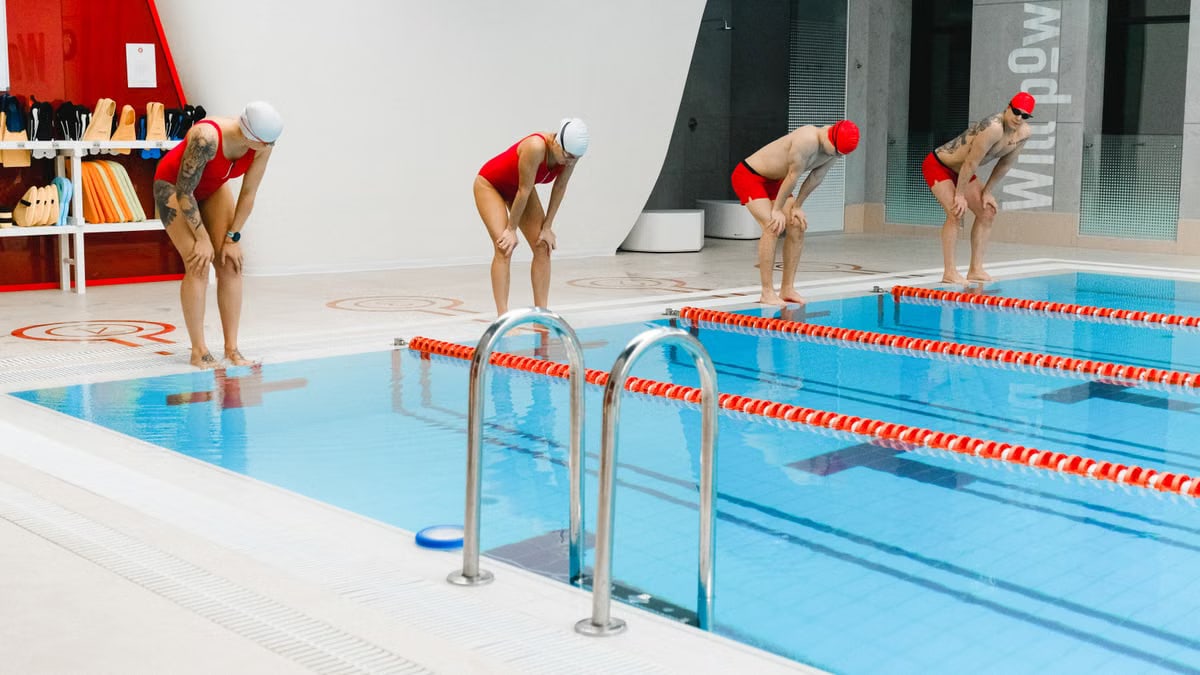 Swimmers in red gear bend before diving into an indoor pool, ready for action.
